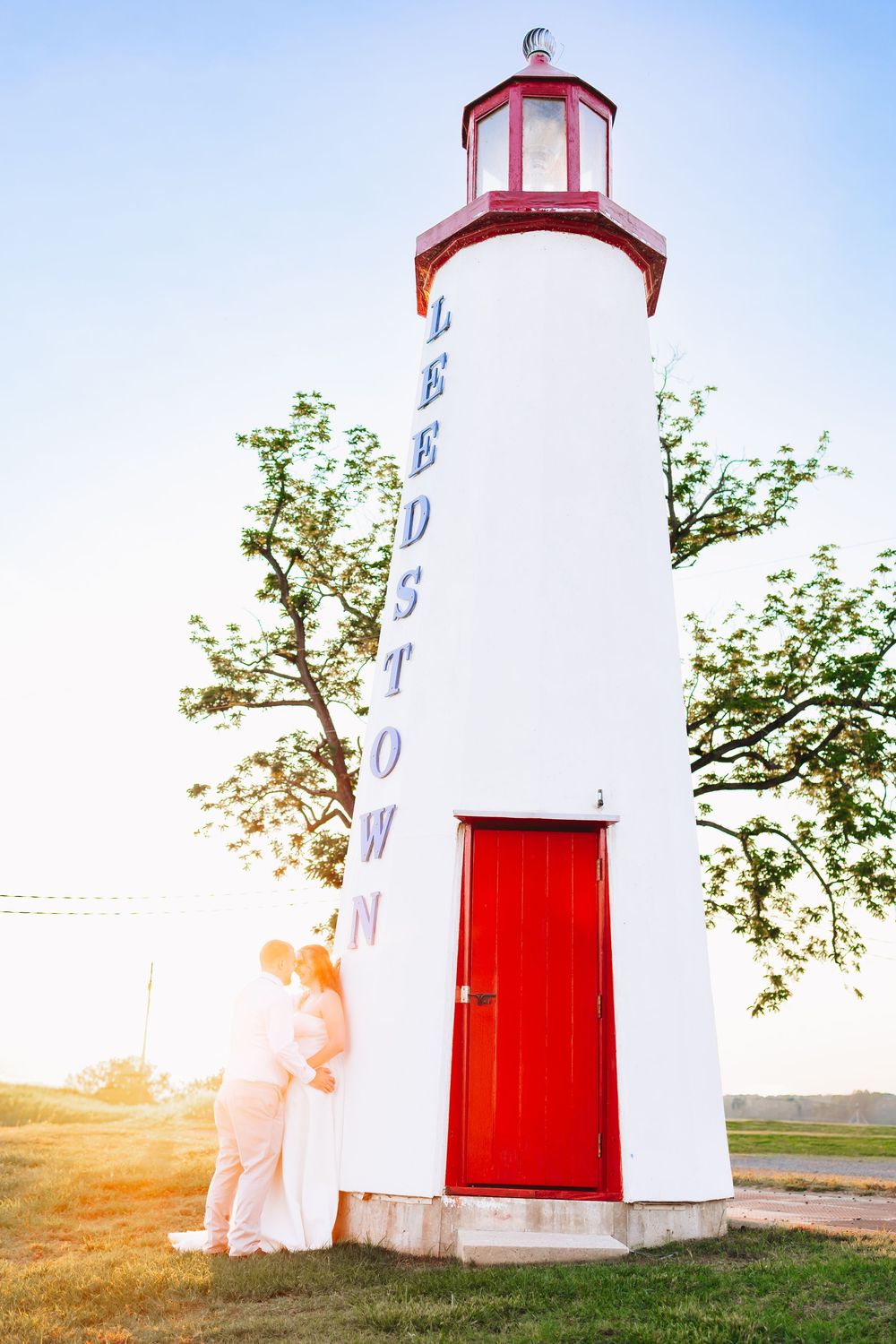 Couple embracing by the Ledstown lighthouse with a red door, surrounded by green trees at golden hour.
