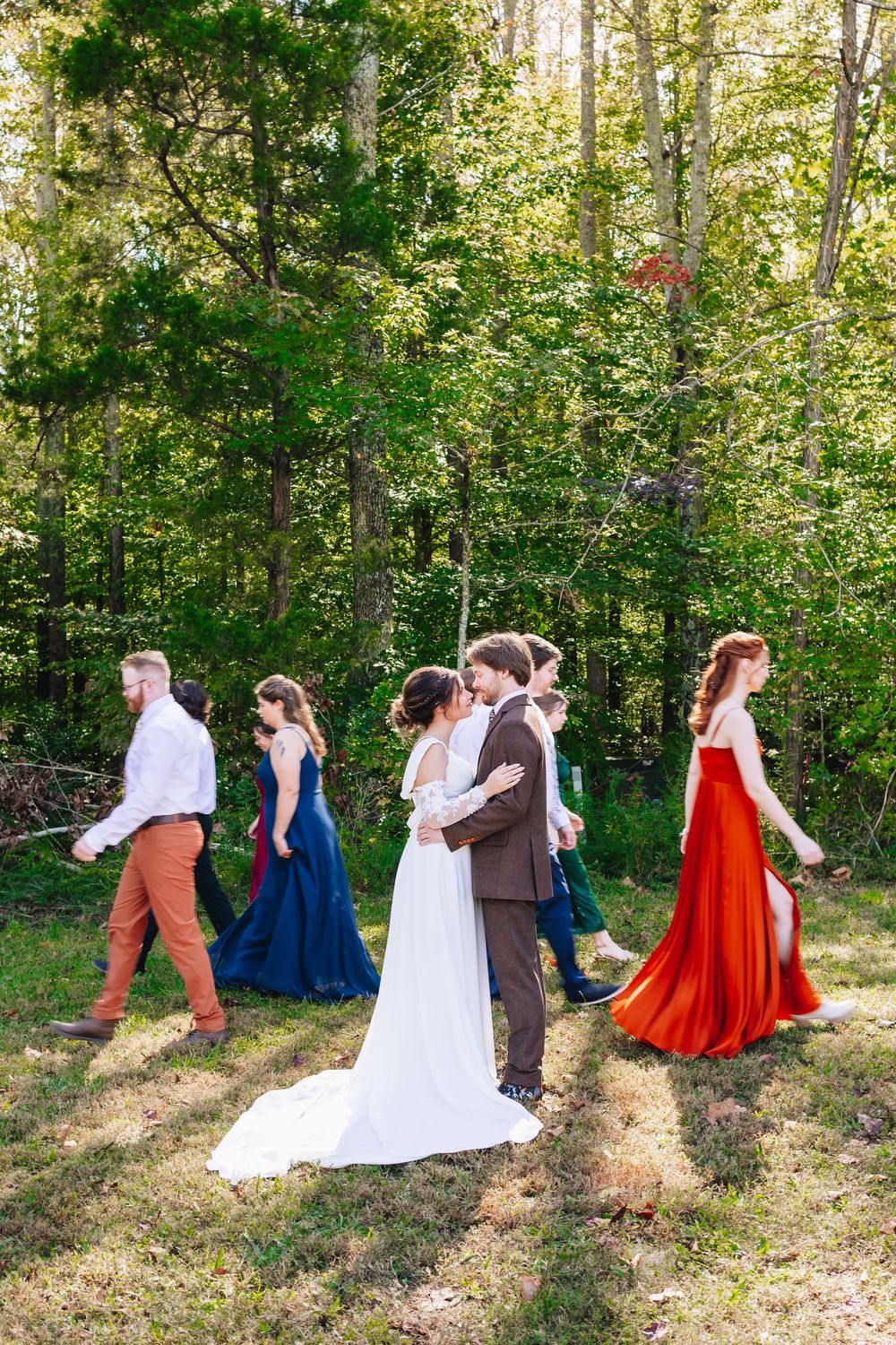 Bride and groom share a kiss surrounded by wedding party in colorful gowns outdoors in a lush green forest setting.