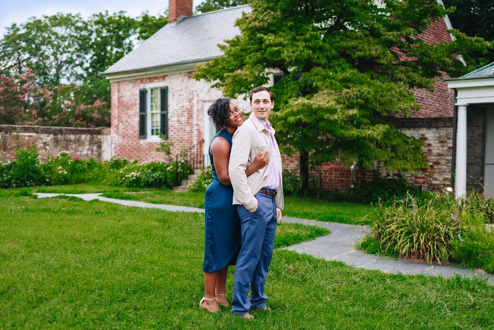 Couple posing together in lush green garden outside historic brick building, woman in blue dress embracing man in white shirt.
