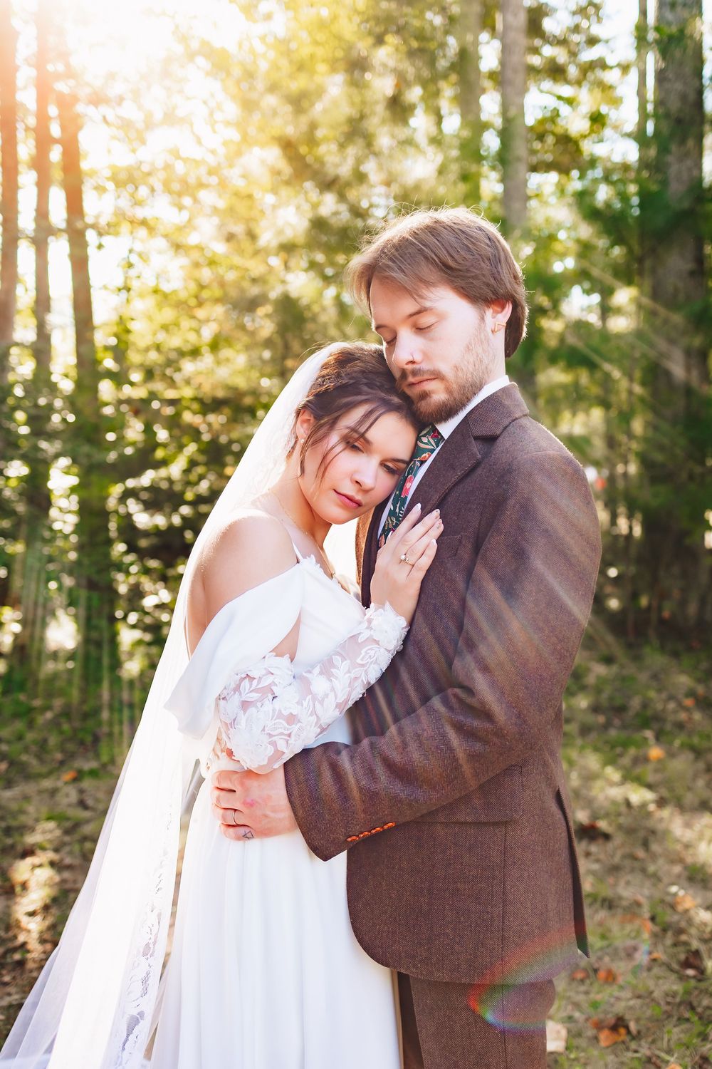 Bride in white lace gown and groom in brown suit embrace tenderly in sunlit forest during outdoor wedding photo session.