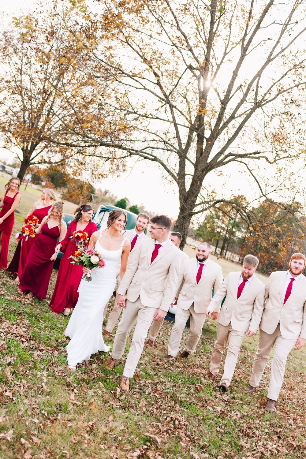 Wedding party walks outdoors in fall, bridesmaids in red gowns and groomsmen in tan suits with red ties.