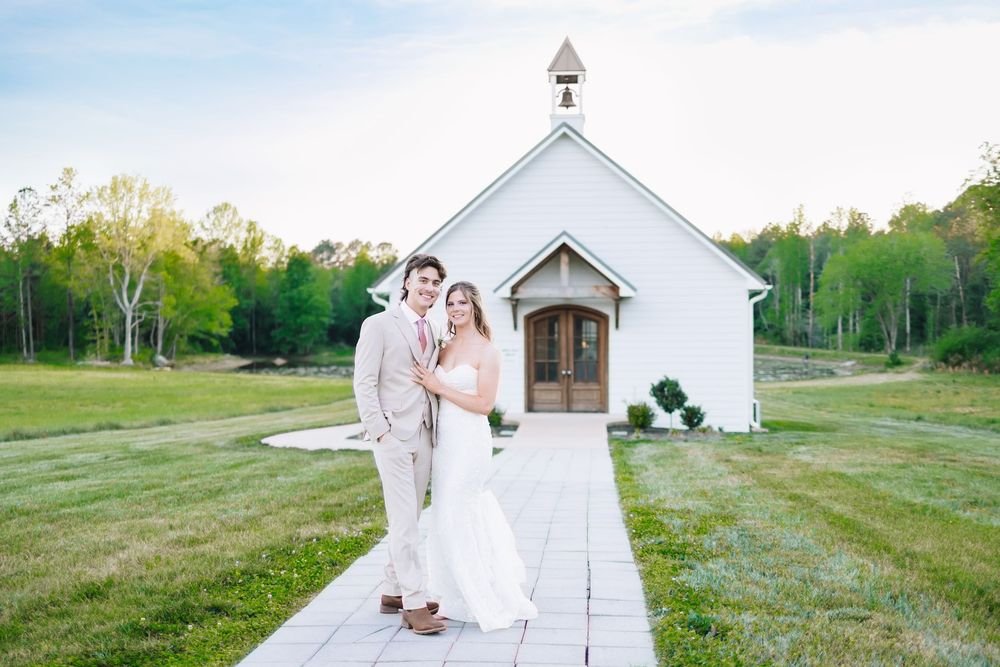 Bride and groom posing together on a stone path in front of a charming white chapel surrounded by lush greenery.