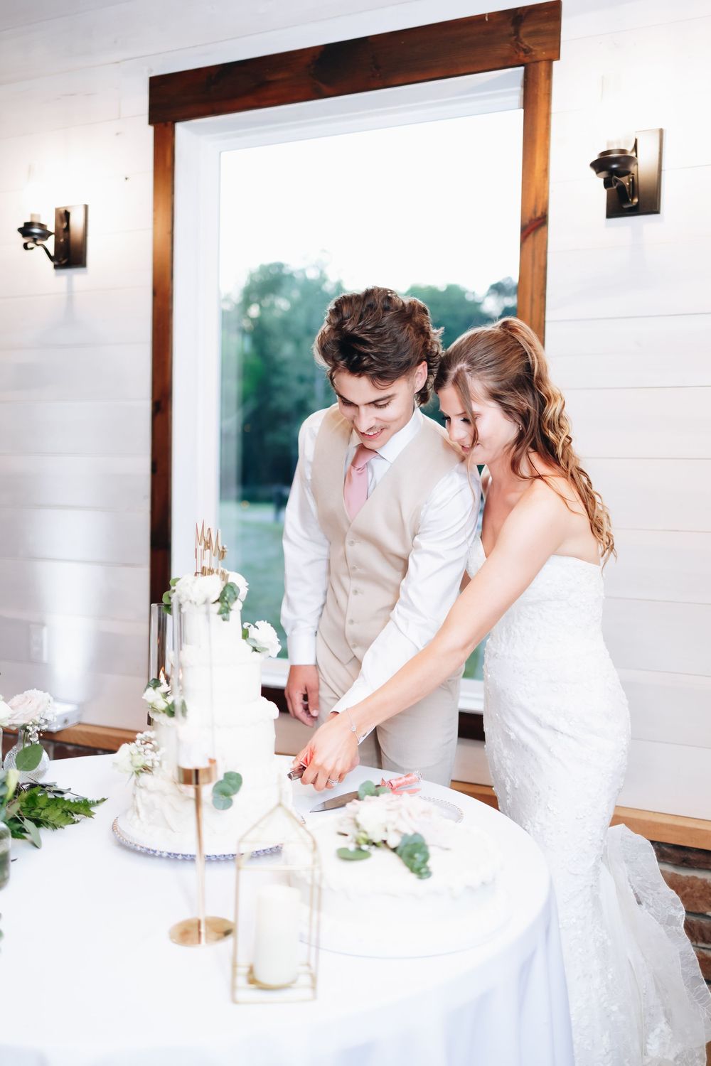 Bride and groom cutting their white wedding cake together at a rustic indoor reception venue.