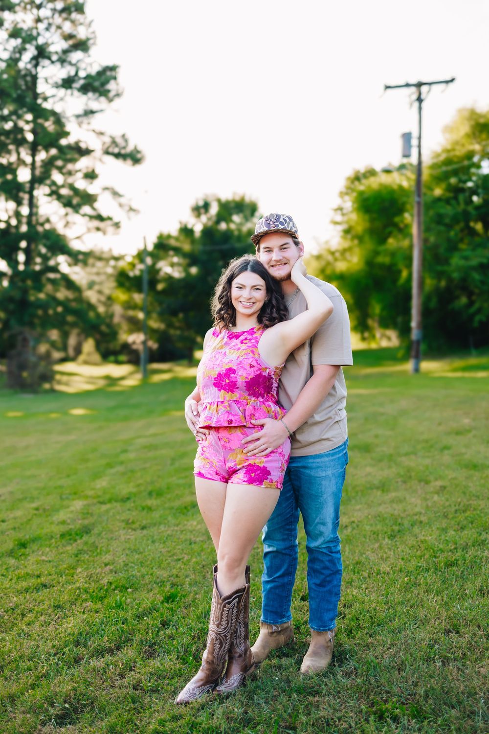 A young couple embraces outdoors on green grass, she wears a pink floral romper with cowboy boots, he wears jeans and a cap.