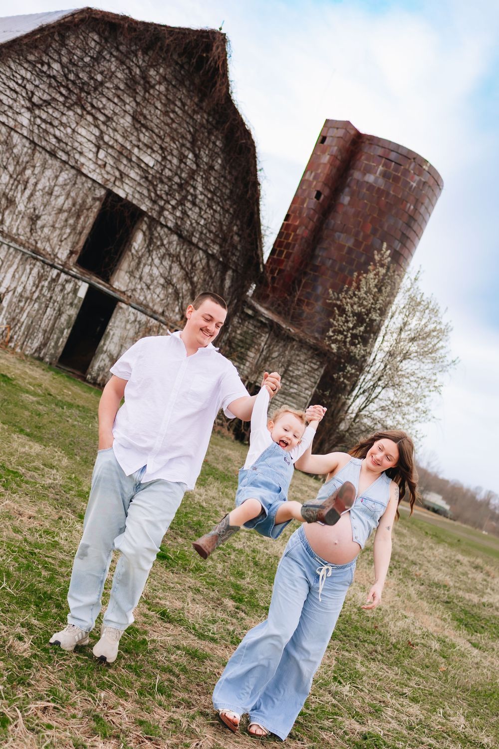 Happy family swinging toddler outdoors near rustic barn and silo, pregnant mother in blue jumpsuit.