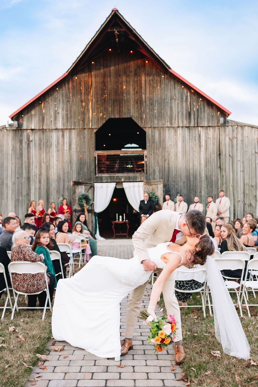 Groom dips bride for a kiss at outdoor barn wedding ceremony while guests watch from white chairs.