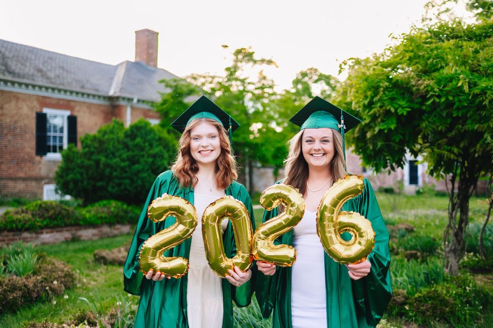 Two smiling graduates in green caps and gowns hold gold 2026 balloon numbers outdoors near a brick building.