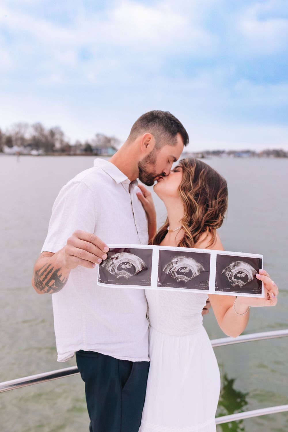 Couple kissing on waterfront dock while holding ultrasound photos, announcing pregnancy in a romantic outdoor setting.