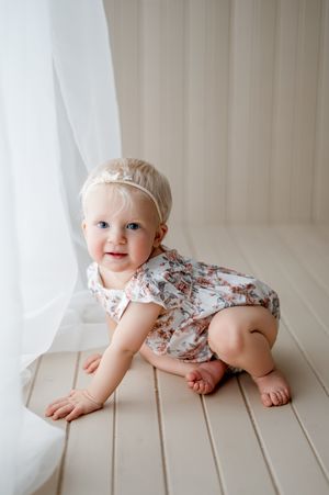 Baby in floral romper crawls playfully on white wooden floor near sheer curtains.