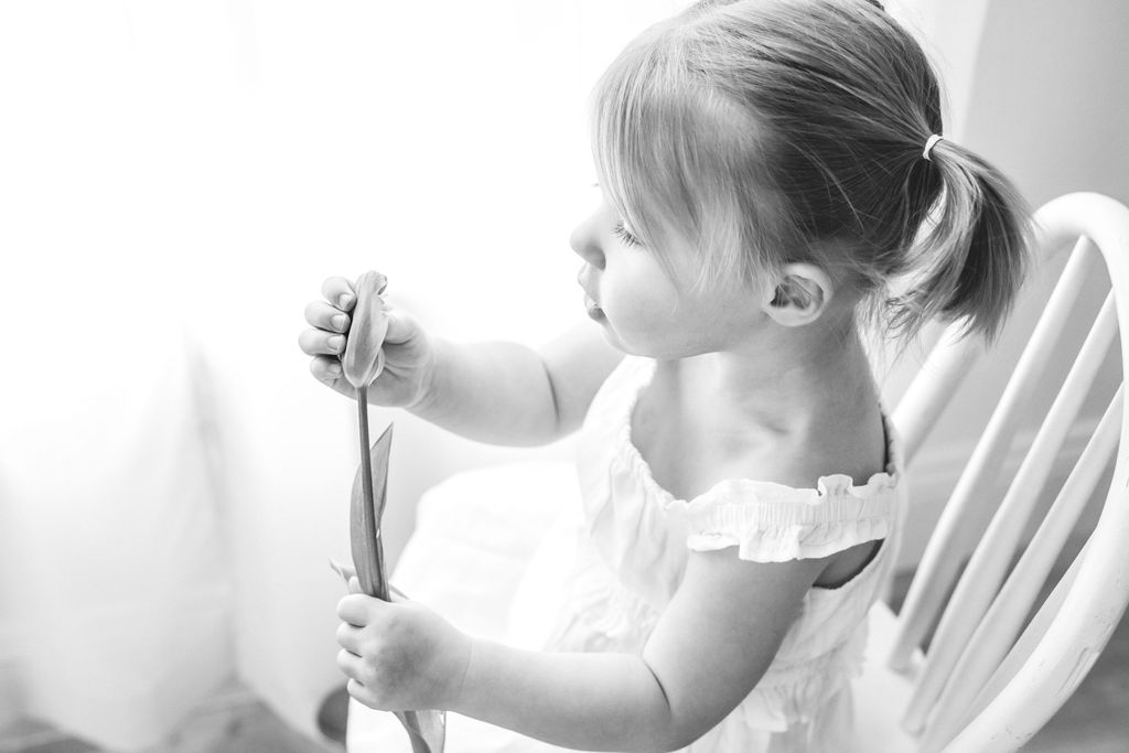 Black and white photo of a young girl with pigtails sitting on a chair, holding a spoon.