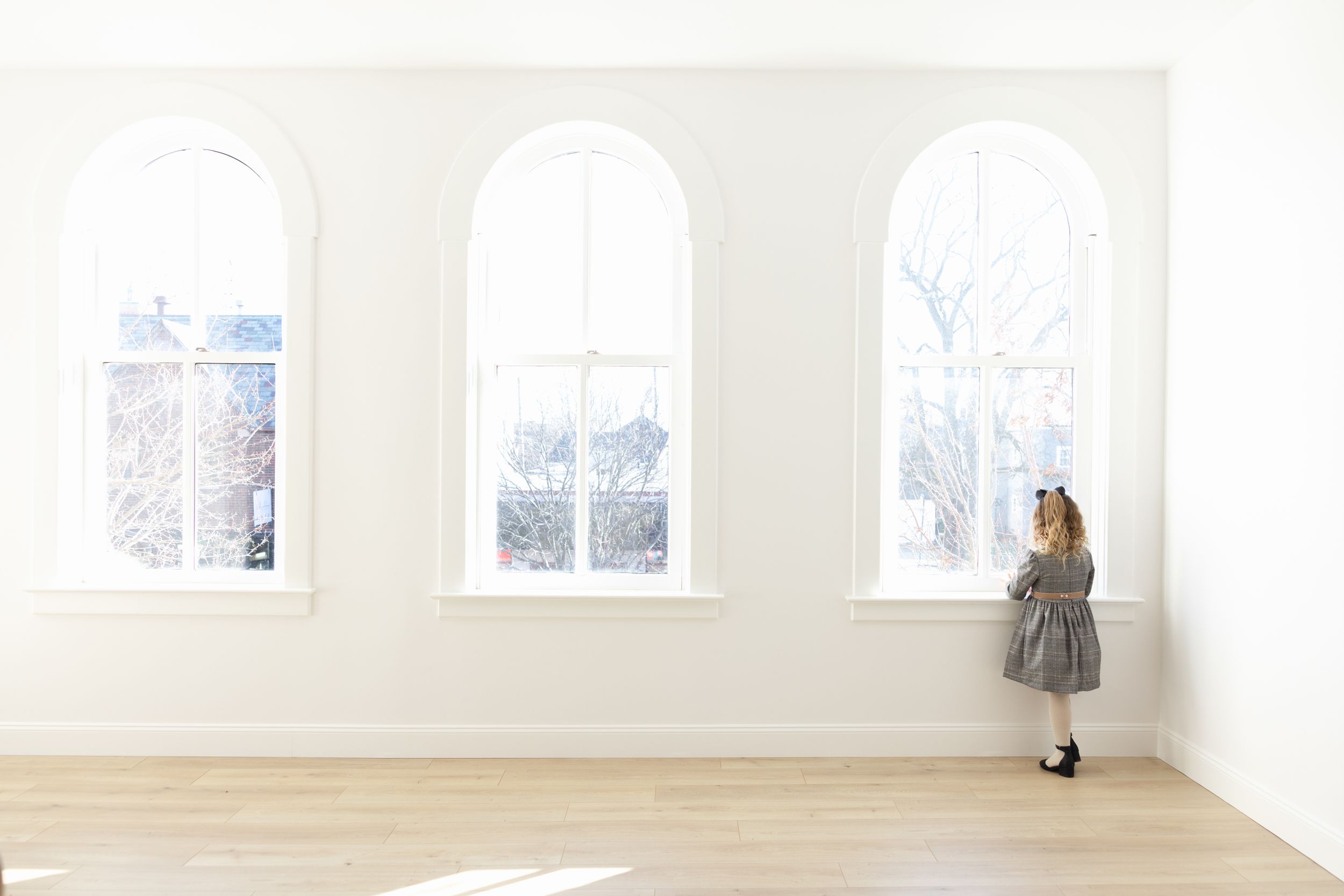 A young girl in a gray dress stands looking out three tall arched windows in a bright, minimalist white room with wood floors.