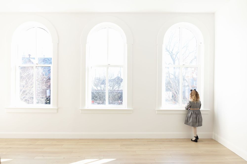A young girl in a gray dress stands looking out three tall arched windows in a bright, minimalist white room with wood floors.