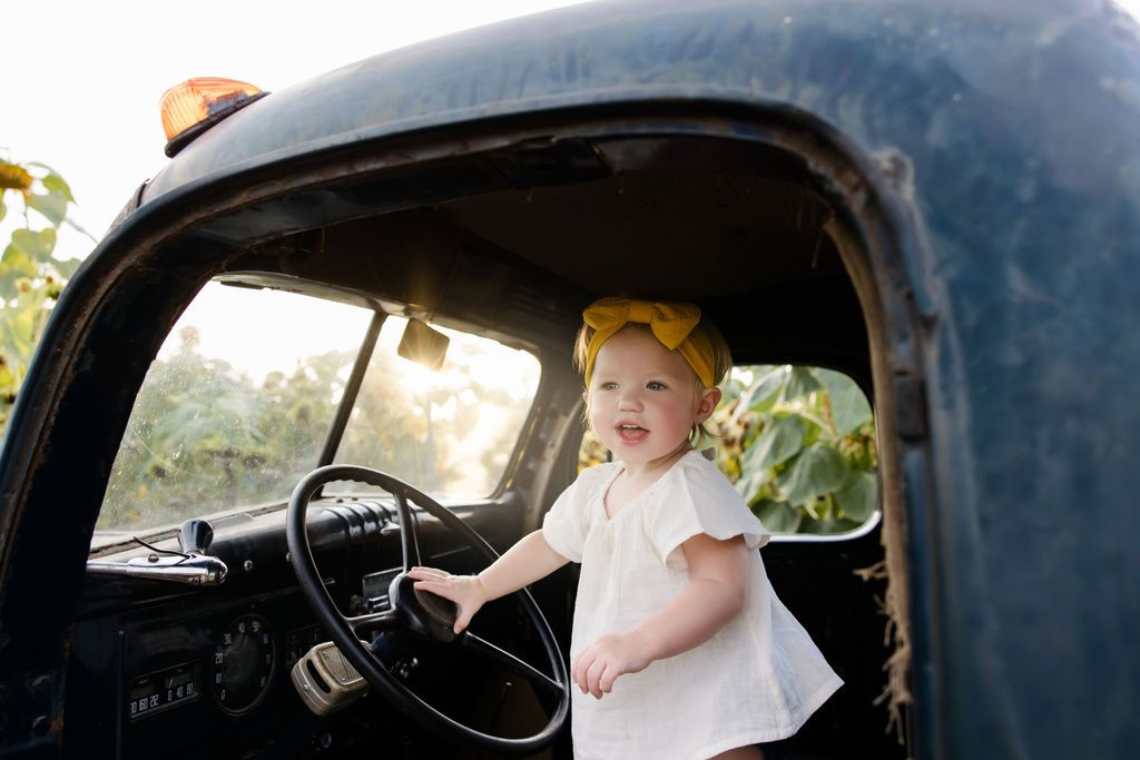 Toddler girl in white dress and yellow bow stands at steering wheel of vintage black truck outdoors.