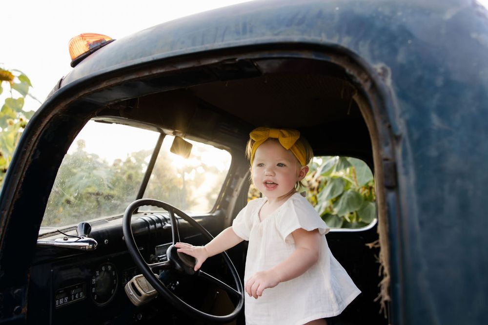 Toddler girl in white dress and yellow bow stands at steering wheel of vintage black truck outdoors.