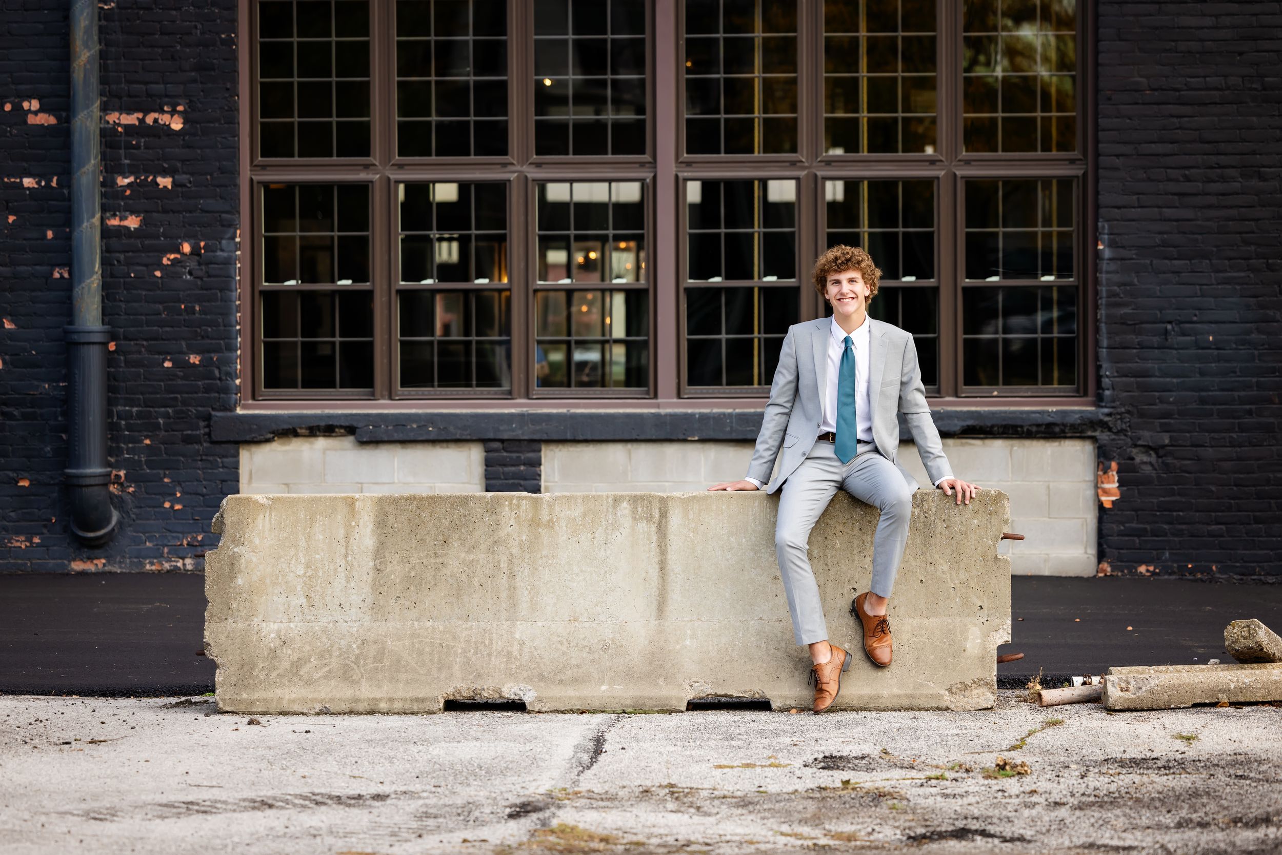 Young man in light gray suit and green tie sitting on concrete barrier outside industrial brick building.