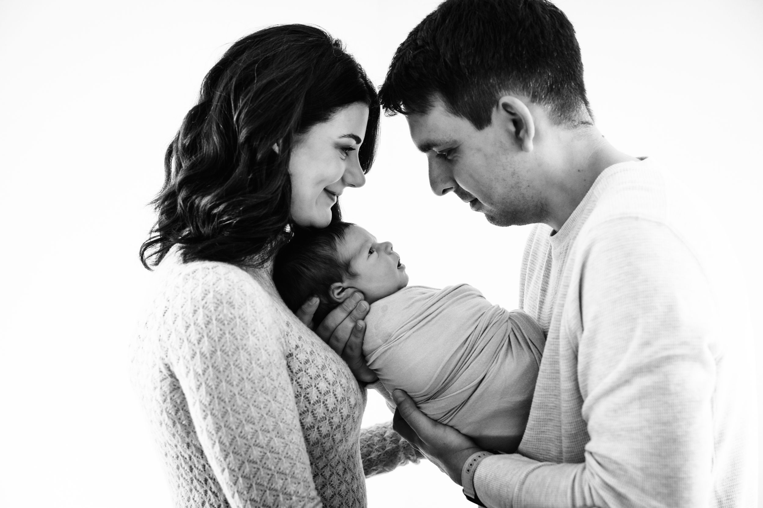 Black and white photo of smiling parents tenderly holding their swaddled newborn baby together.