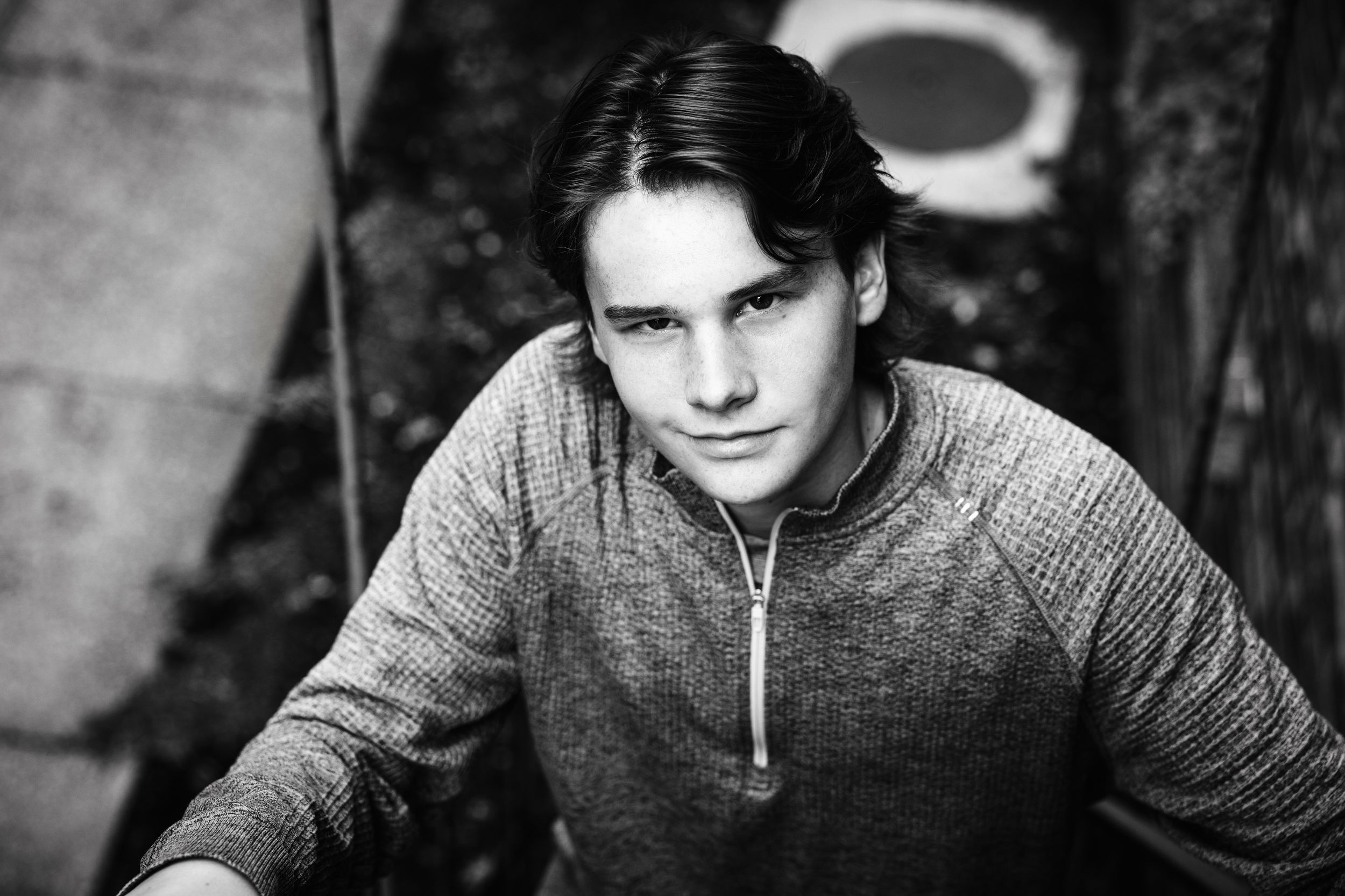 Black and white portrait of a young man in a gray zip-up sweater with dark hair, looking directly at the camera.