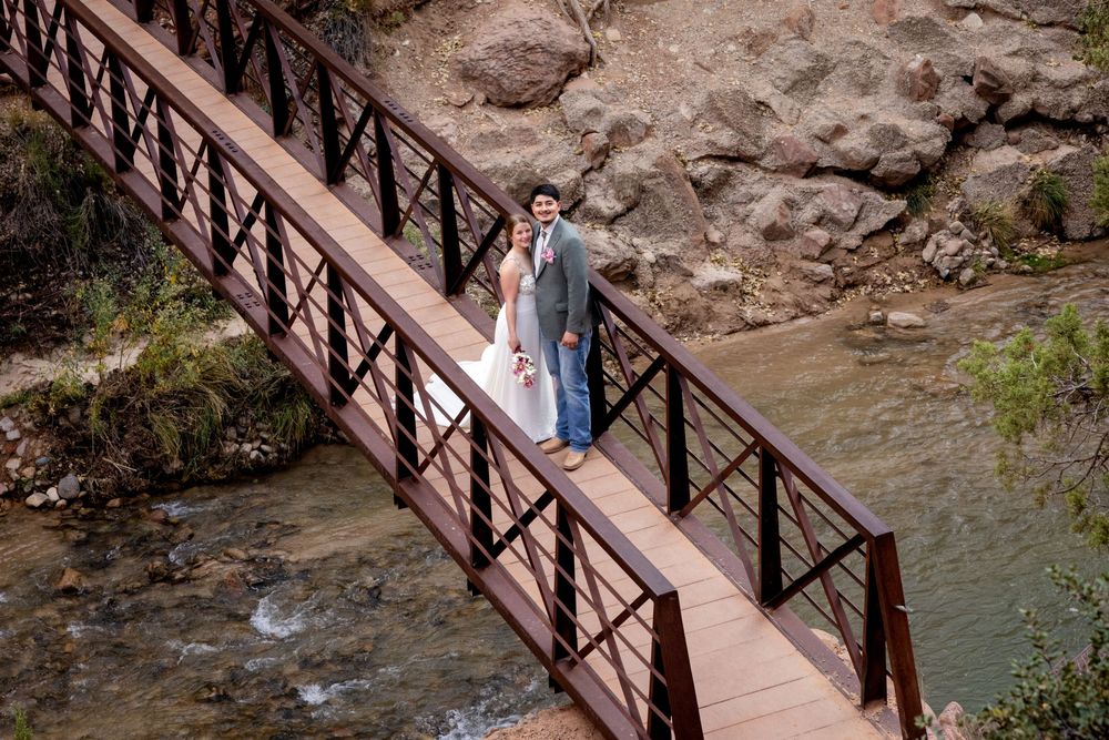 Bride and groom pose on a wooden bridge over a rocky stream in a scenic outdoor wedding photo.