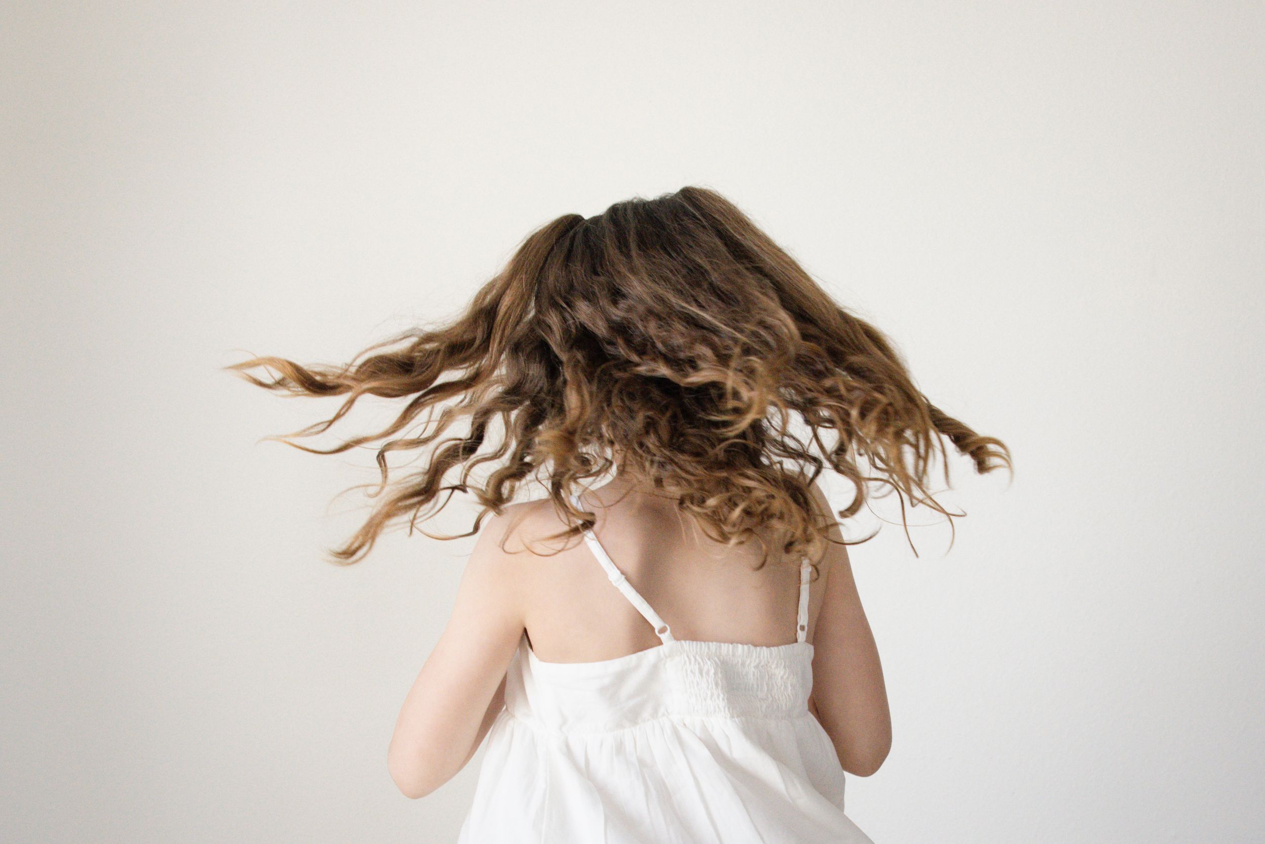 A young girl in a white dress tosses her long curly brown hair against a light background.