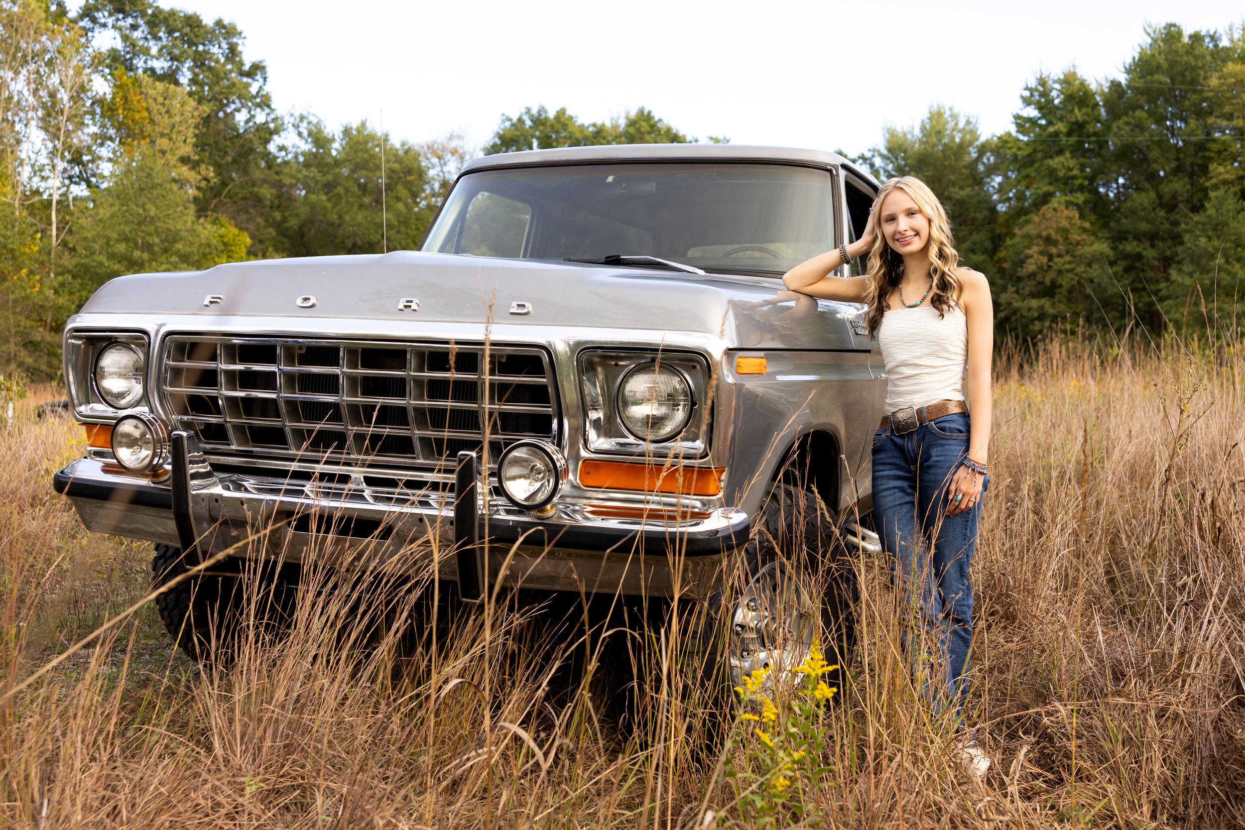 Young blonde woman posing beside a classic silver Ford pickup truck in a rural field with autumn foliage.