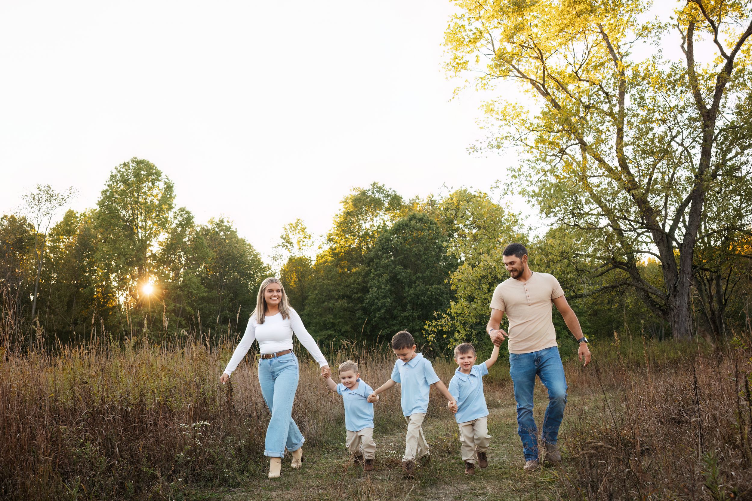 Family of five walking hand-in-hand through a sunlit meadow during golden hour in autumn.