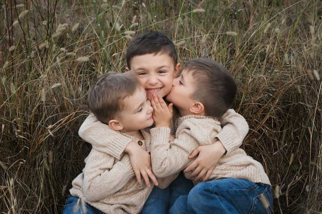 Three young boys in beige sweaters share kisses and hugs outdoors in a field of tall golden grass.