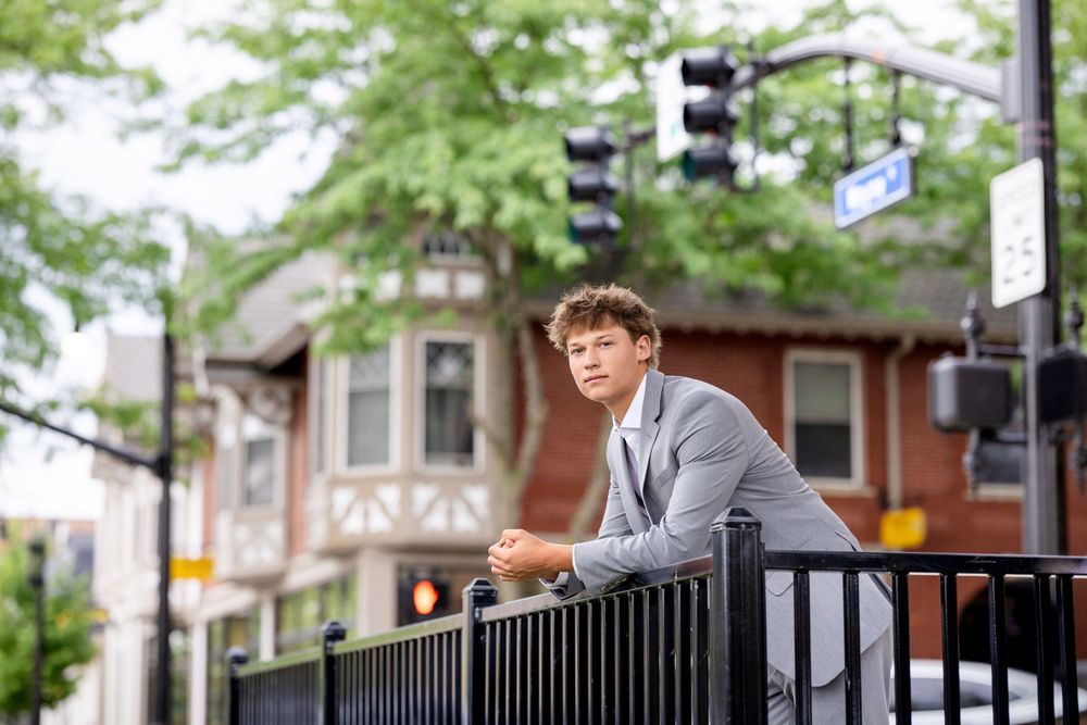 Young man in gray blazer leaning on black iron fence on urban street with green trees and buildings.