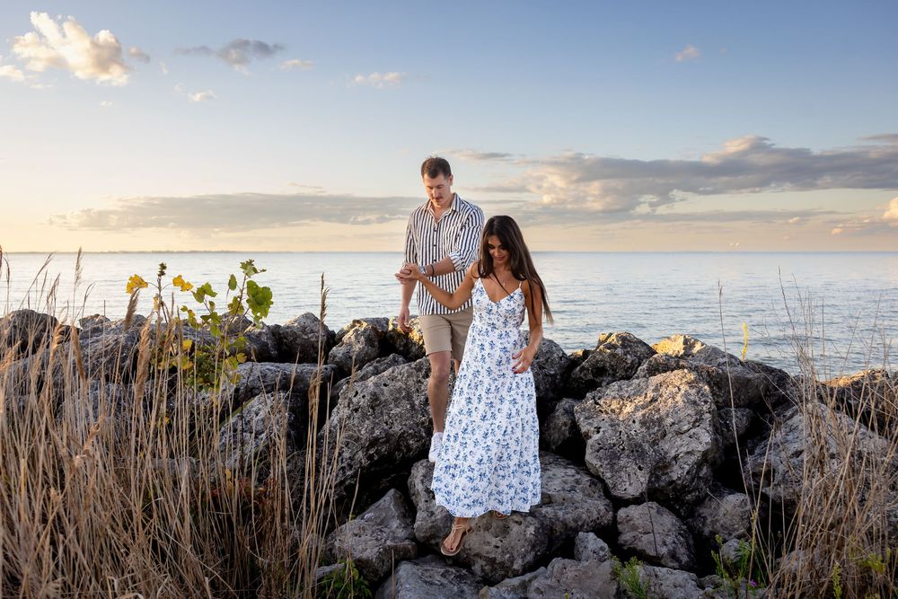 Couple walking on rocky shoreline at sunset, woman in floral dress and man in casual attire with ocean backdrop.
