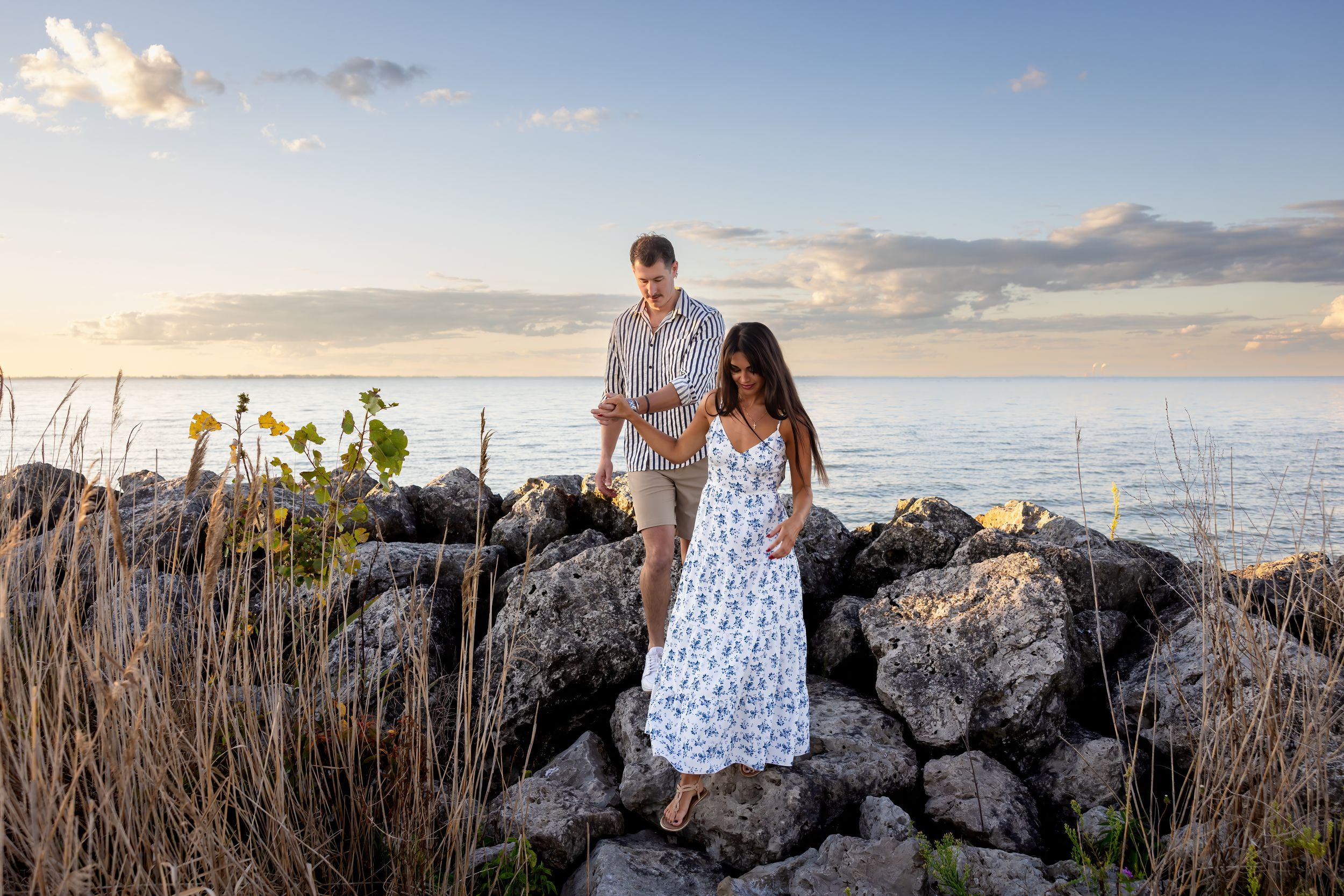 Couple walking on rocky shoreline at sunset, woman in floral dress and man in casual attire with ocean backdrop.