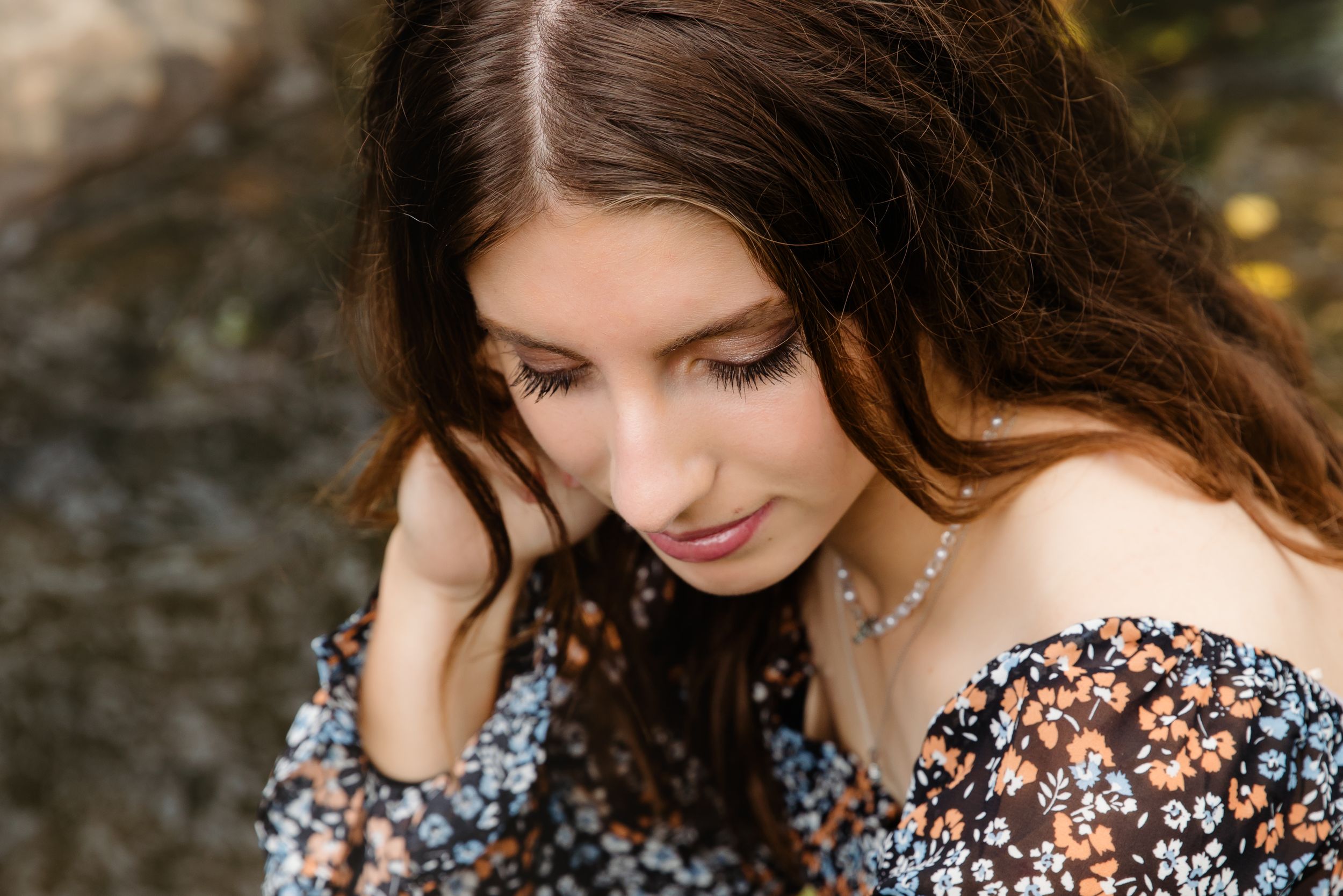 Young woman with long brown hair in floral off-shoulder dress looks down thoughtfully in an outdoor portrait.