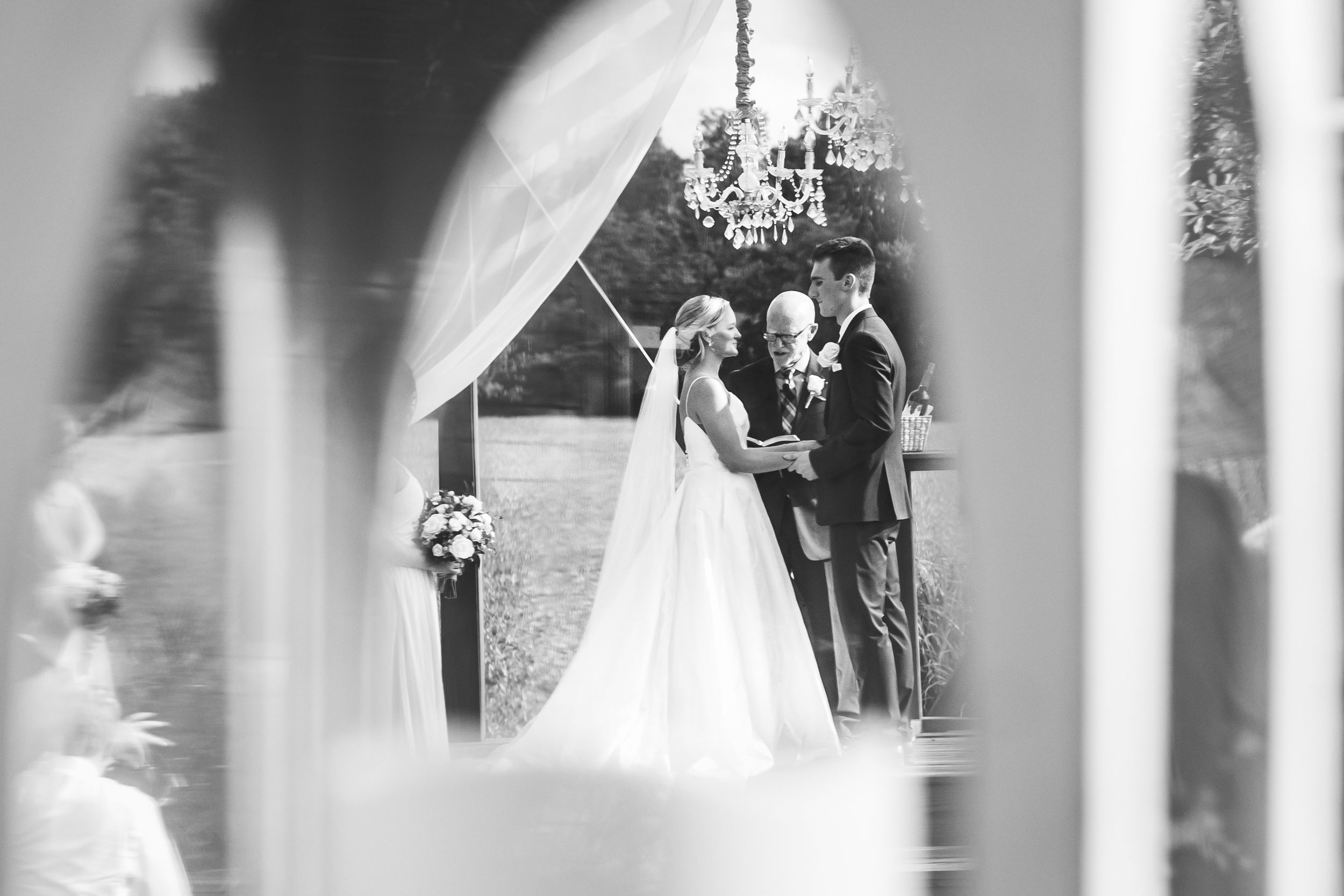Black and white photo of a bride and groom exchanging vows outdoors under a chandelier arch at an elegant wedding ceremony.