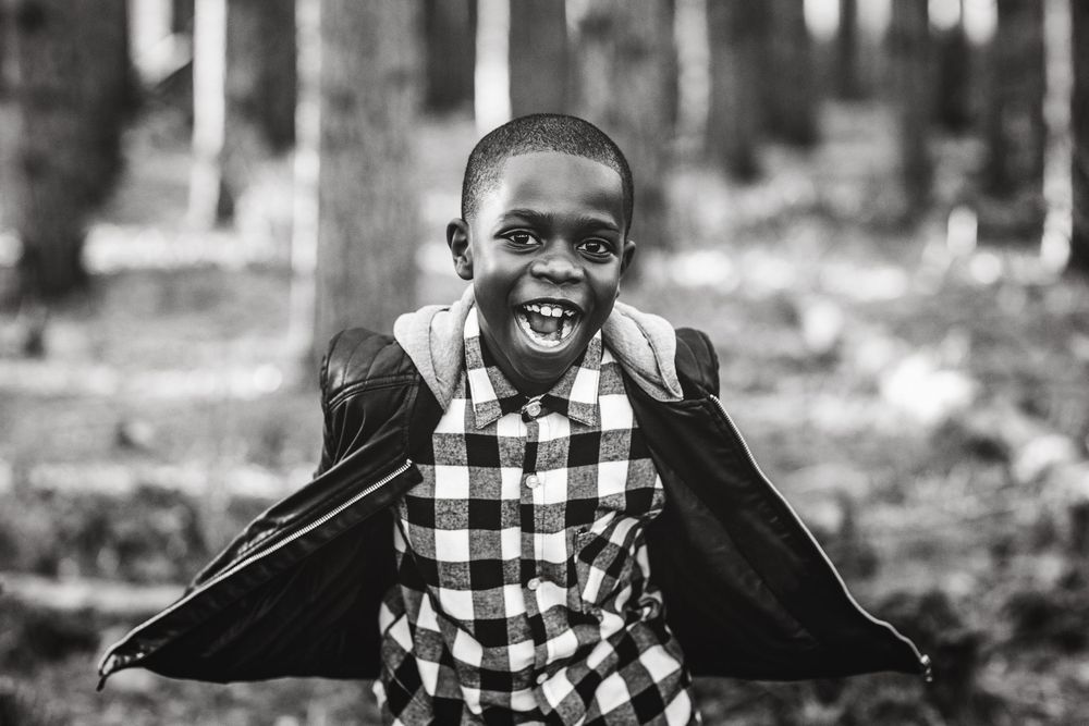 Black and white photo of a joyful young boy in a plaid shirt and cape smiling in a forest setting.