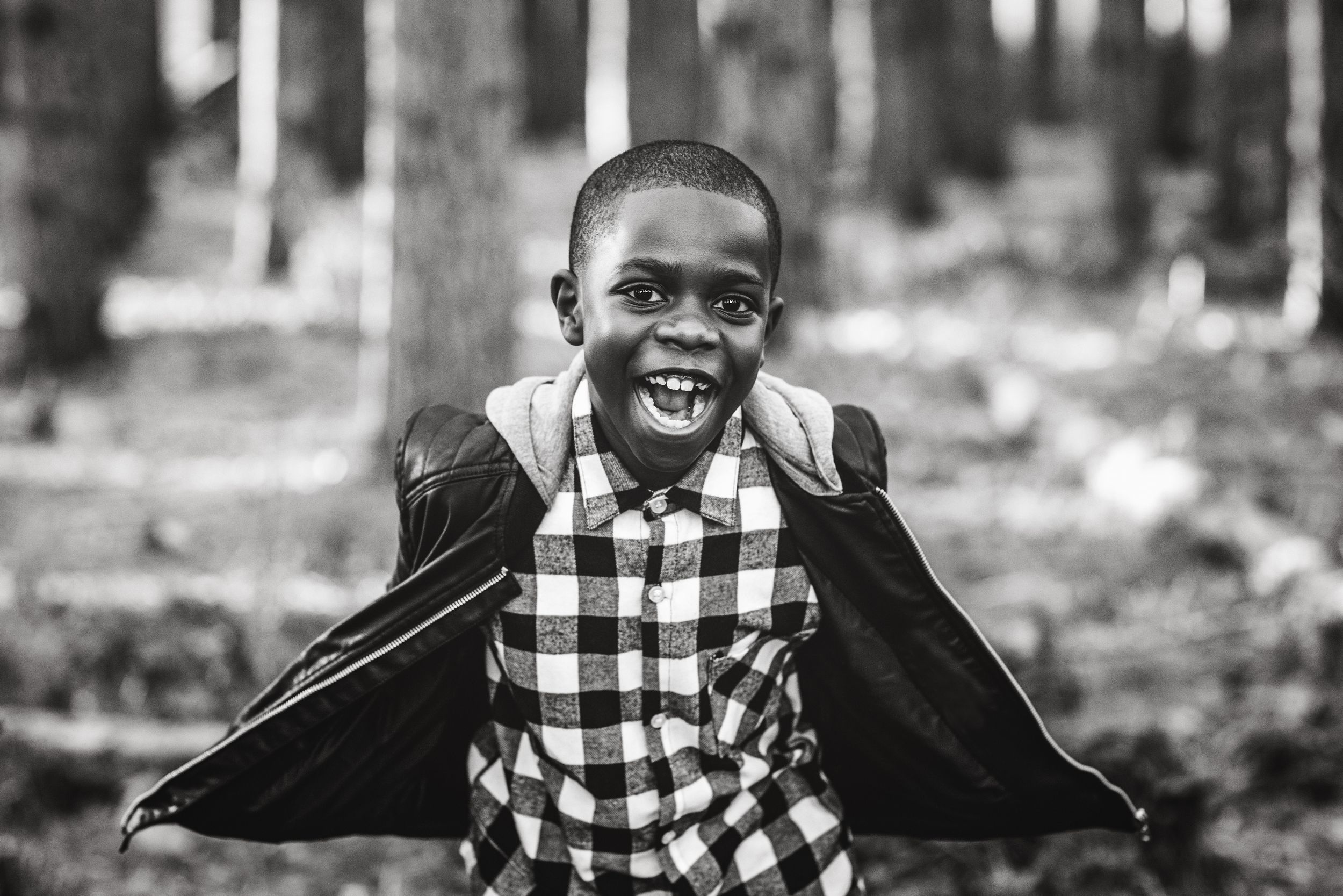 Black and white photo of a joyful young boy in a plaid shirt and cape smiling in a forest setting.