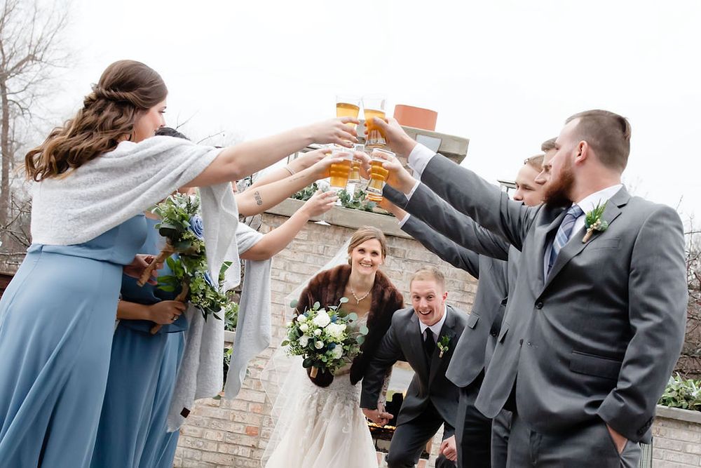 Wedding party toasting with beer glasses outdoors, bride holding bouquet in white gown, bridesmaids in blue, groomsmen in gray suits.