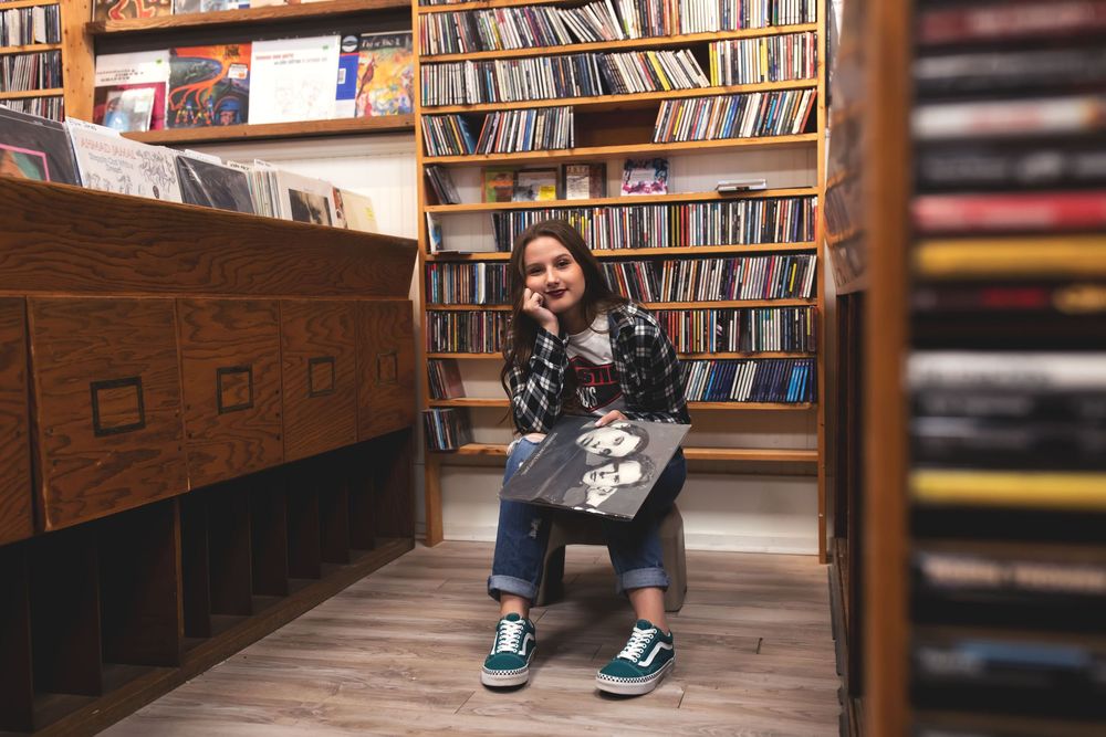 Young woman smiling in a record store, holding a vinyl album, surrounded by shelves of records and CDs.