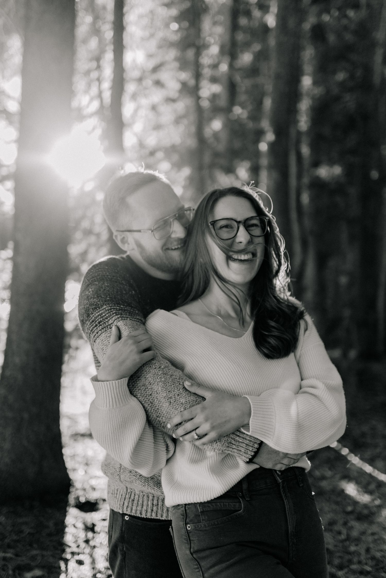 Black and white photo of a laughing couple embracing in a sunlit forest, both wearing glasses and cozy sweaters.