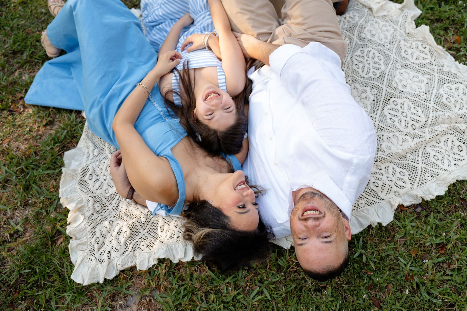 Three people lying on a blanket on grass, smiling and laughing together during an outdoor picnic.