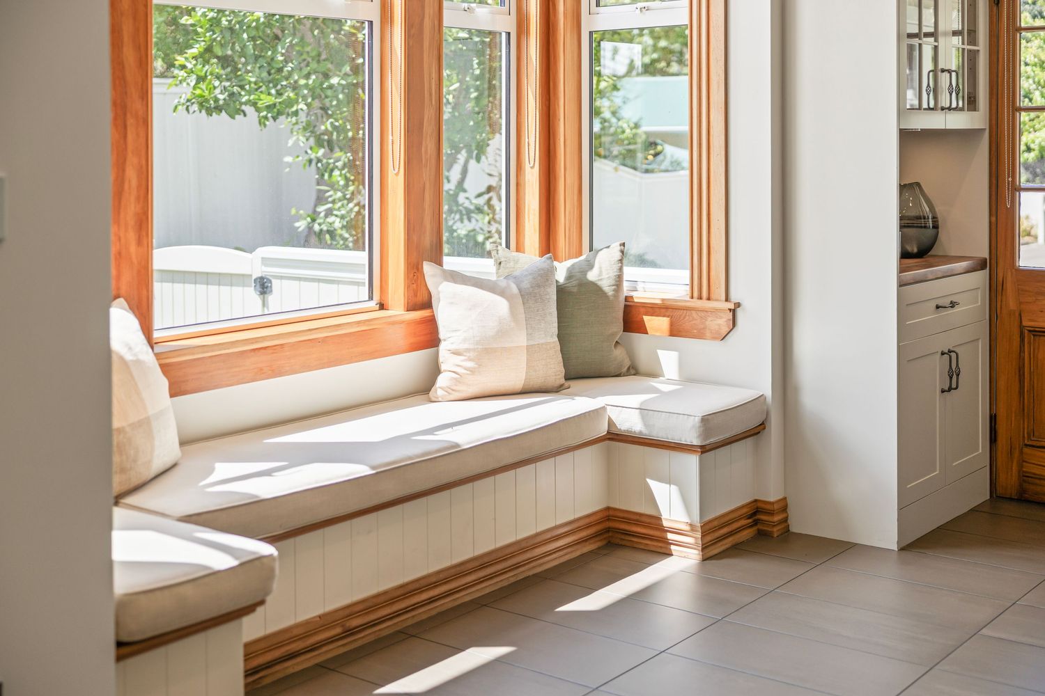 Bright corner window seat with white cushions, green pillows, and wood trim in a sunlit breakfast nook.
