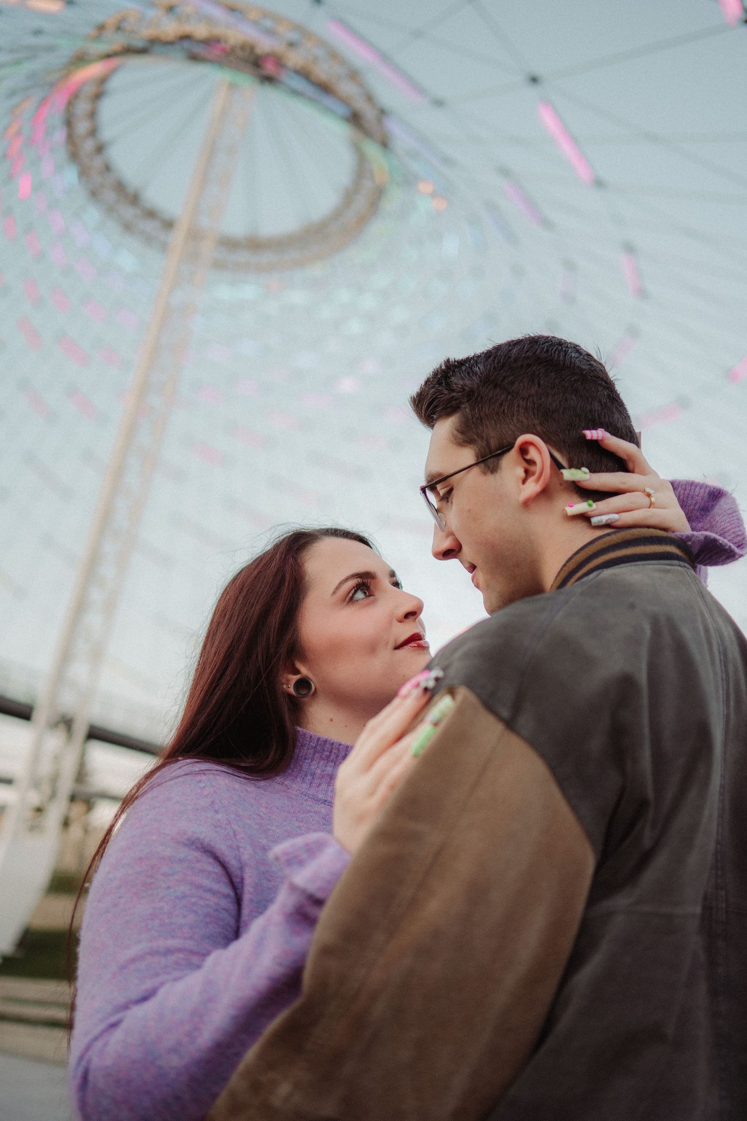 Couple embracing in front of a colorful Ferris wheel, woman in purple sweater gazing at man wearing glasses.