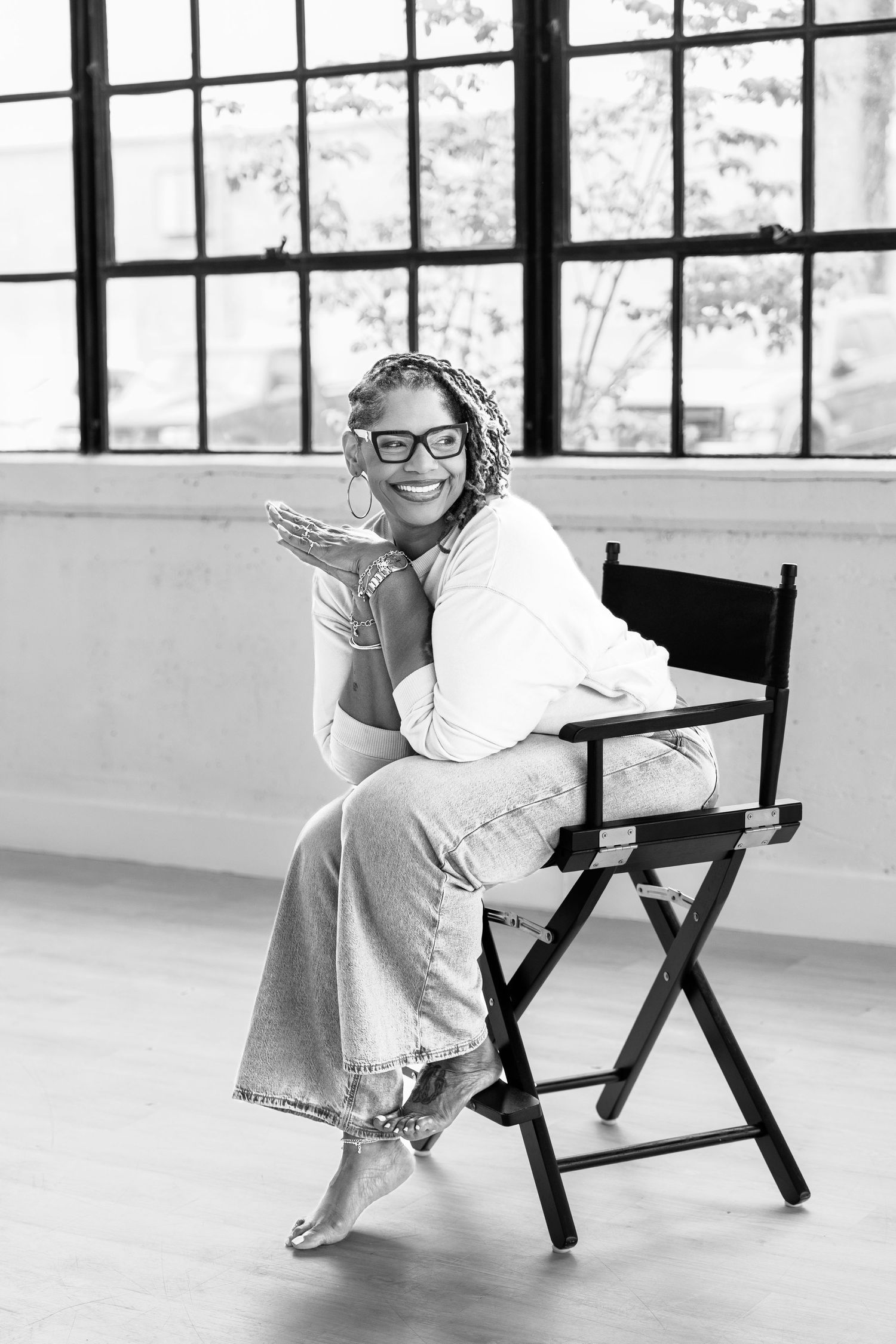 Woman with braids and glasses sits in director's chair, smiling and blowing a kiss in a bright studio.