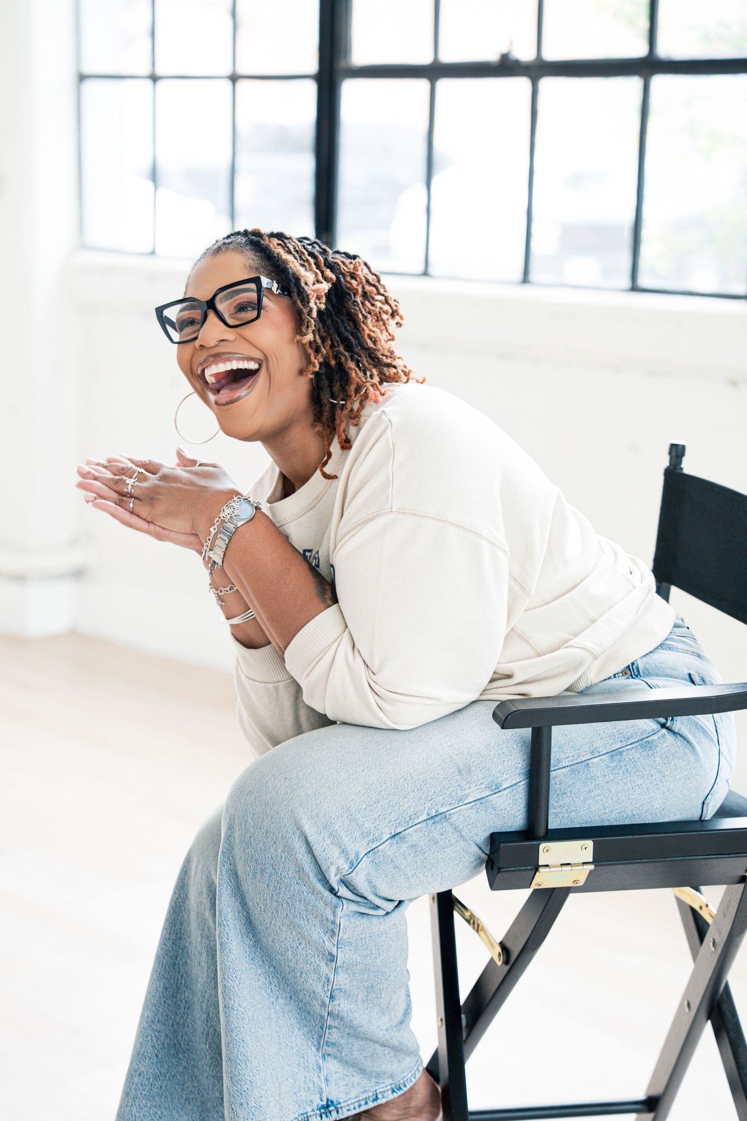 Laughing Black woman with locs and glasses sits in director's chair wearing white sweater and jeans.