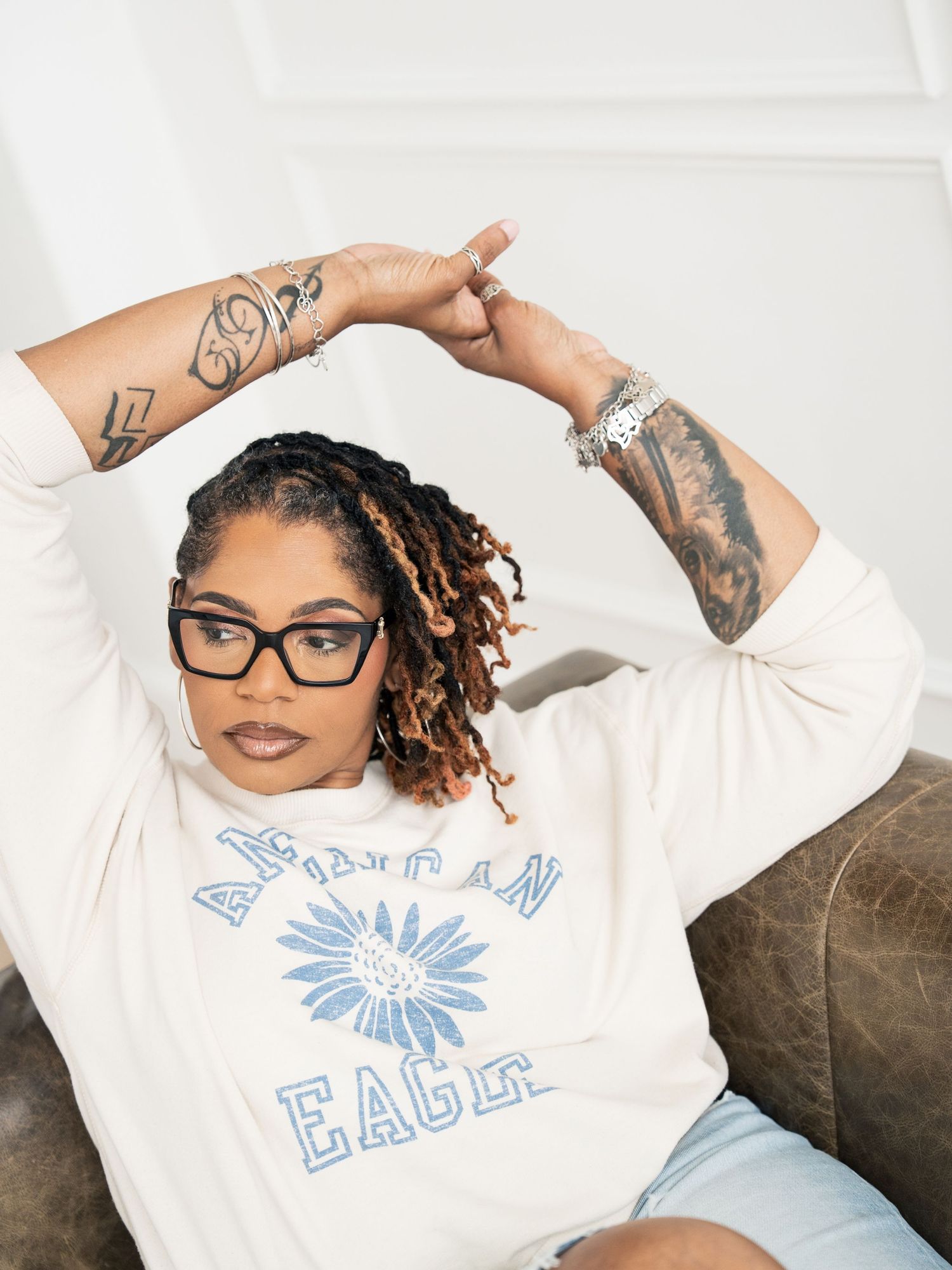 Woman in American Eagle sweatshirt with tattoos, glasses, and locs posing on a couch.