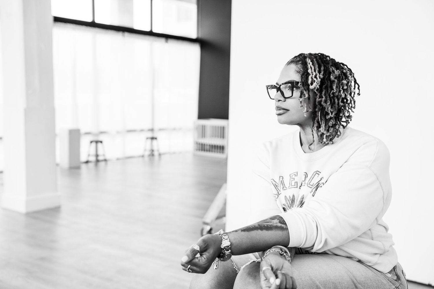 Black and white photo of woman with locs and glasses sitting in bright open space.