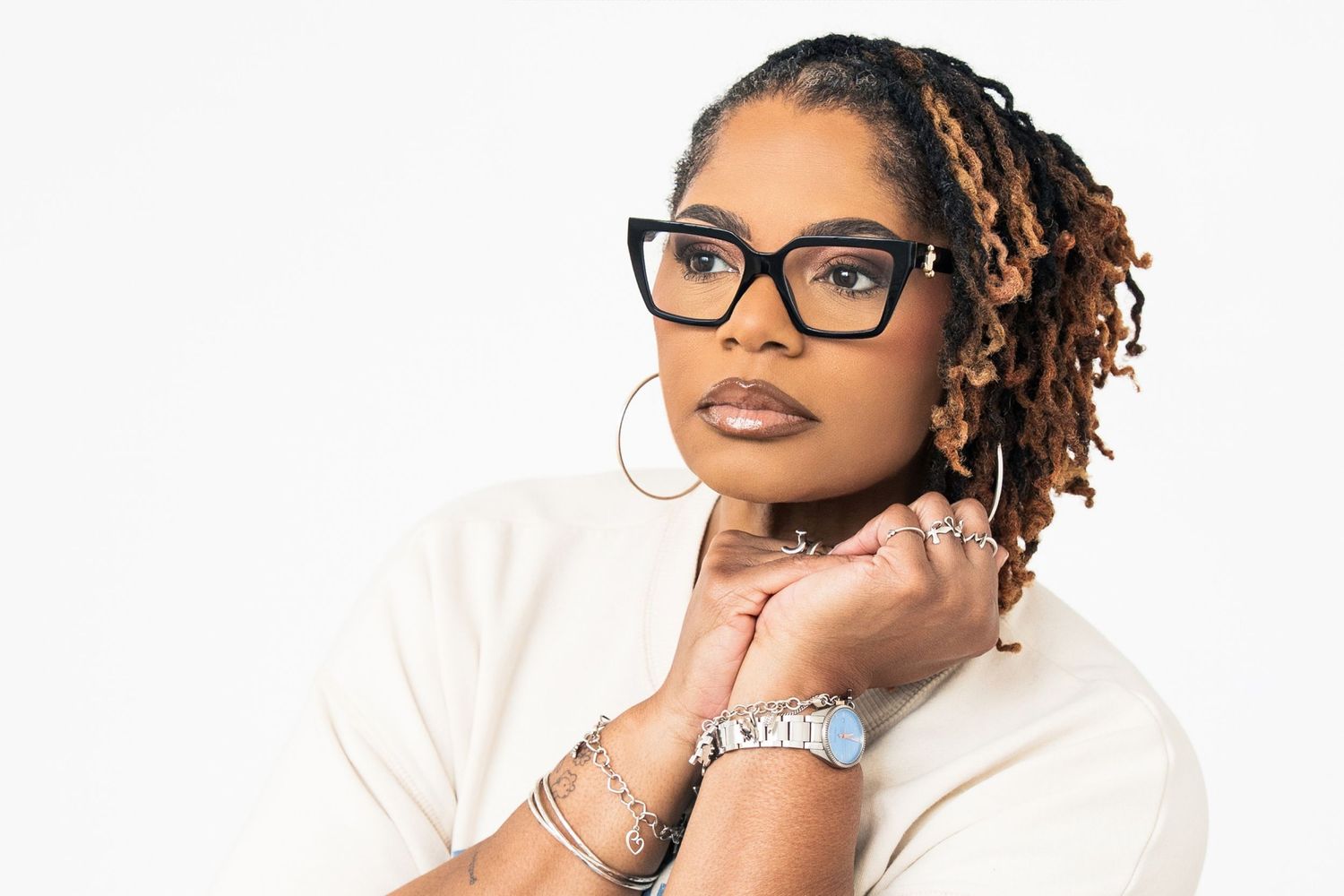 Stylish woman with locs, glasses, hoop earrings, and silver bracelets posing confidently.