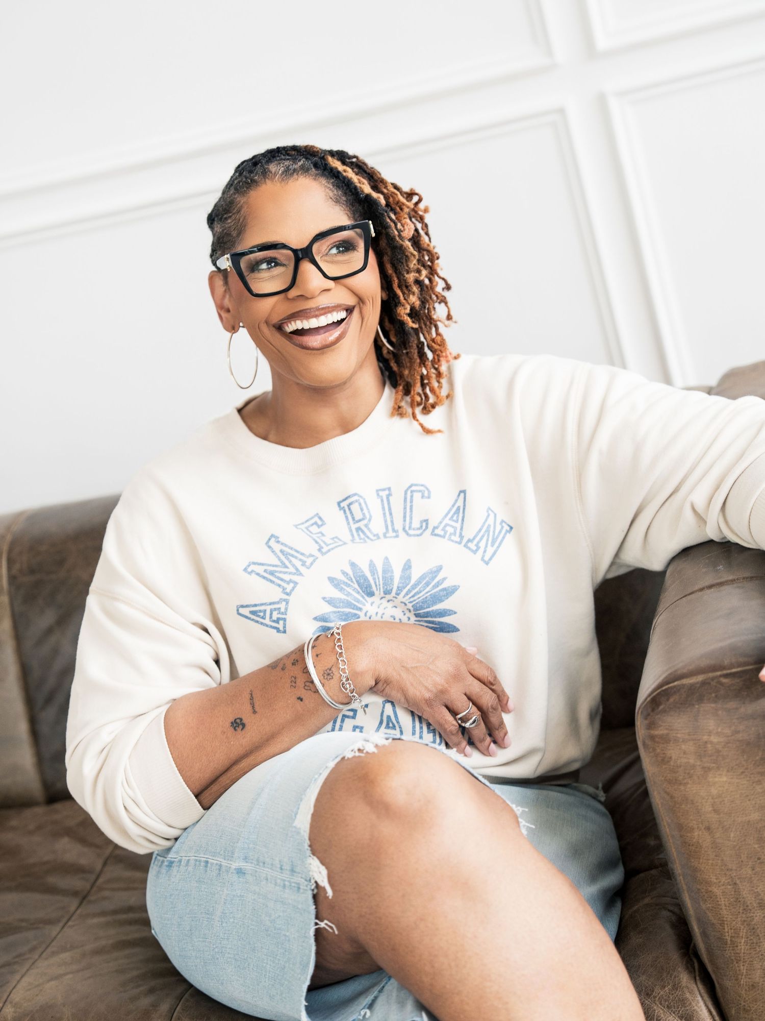 Smiling woman with locs and glasses wearing an American sweatshirt and denim shorts, sitting on a brown leather couch.