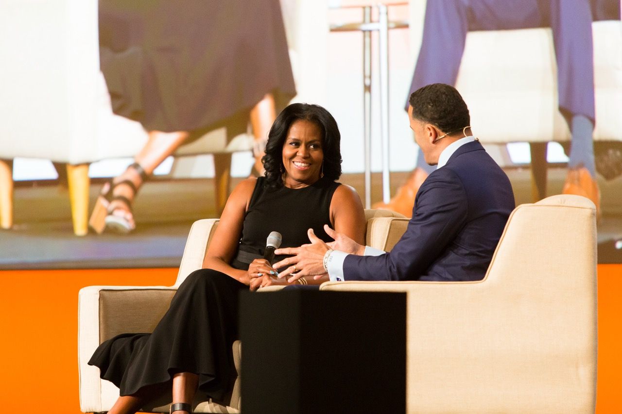 Two people in conversation (One being Michelle Obama) on stage, seated in modern chairs on an orange platform at a professional event.