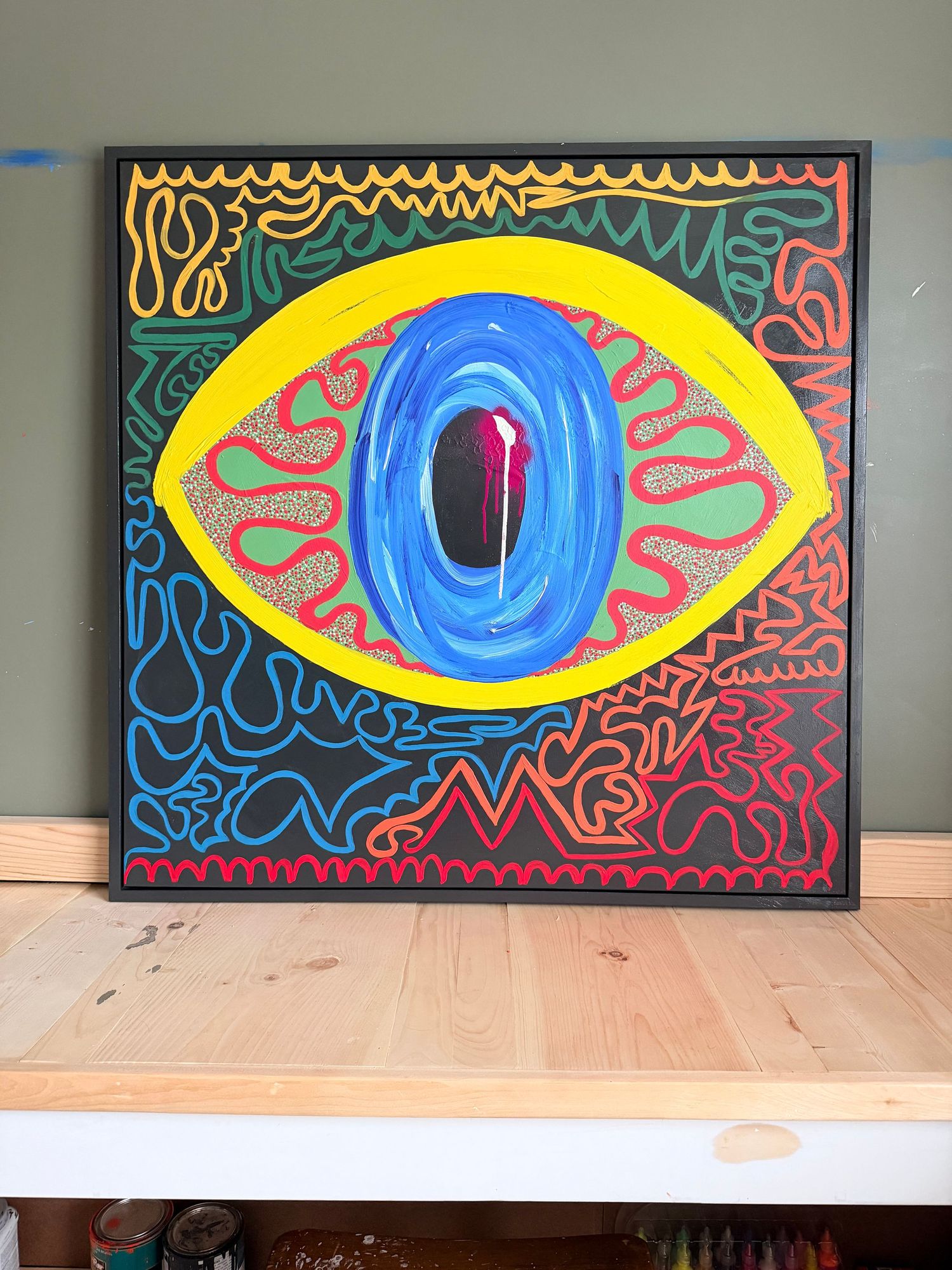 Vibrant psychedelic eye painting on black canvas with yellow, blue, and red abstract patterns in a wooden frame.