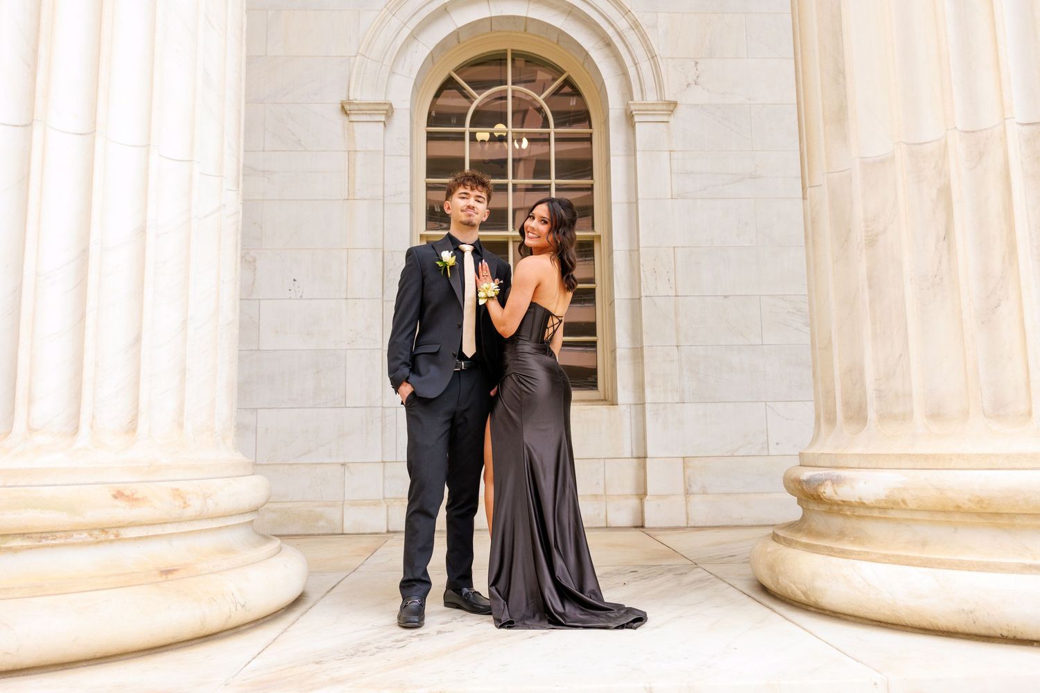 Elegant couple in formal attire posing before grand columns at prom, wearing black tux and gown.