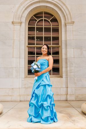 Woman in blue strapless ball gown holding bouquet poses in front of elegant stone arch window.