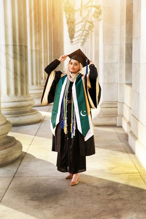 Graduate in black cap and gown with green Pakistani flag sash poses between stone columns in sunlit hallway.